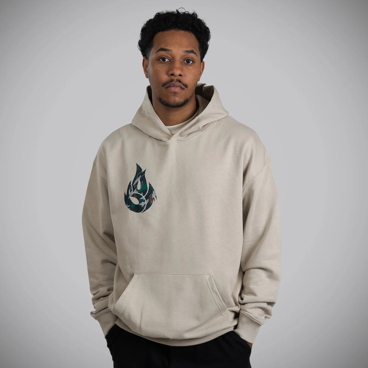 A man wearing a light beige pullover hoodie with a dark teal and black flame graphic on the left chest, standing against a neutral gray background.