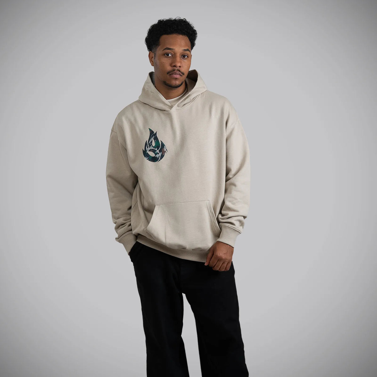 A man wearing a light beige hoodie with a dark blue and white graphic logo on the left chest, paired with black pants, standing against a neutral gray studio background.