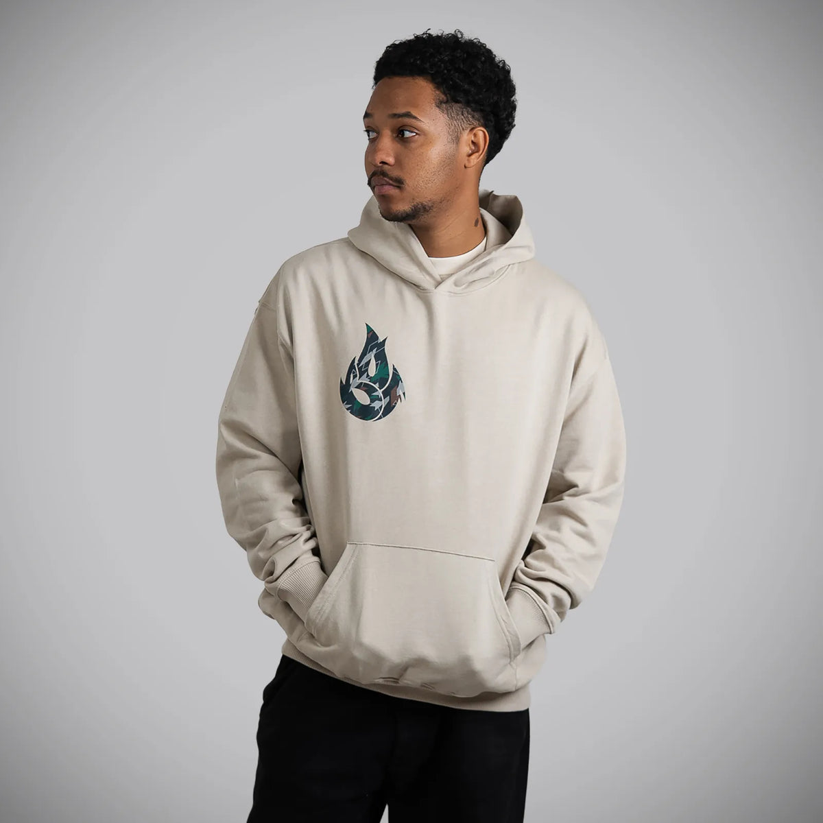 A man wearing a light beige hoodie with a dark teal and white graphic logo on the left chest, paired with black pants, standing against a neutral gray studio background.